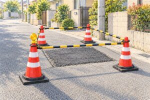 住宅地の道路の小さな舗装補修の場所を、赤いカラーコーン4本とコーンバーで四方から囲んでいる水彩画。手前のコーンの頭に工事灯